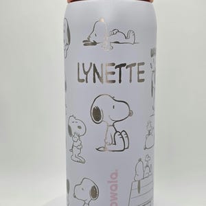 May include: A white insulated water bottle featuring gold Snoopy illustrations and the name "LYNETTE" in gold. The bottle has a light green and orange lid and the brand name "owala" in pink.