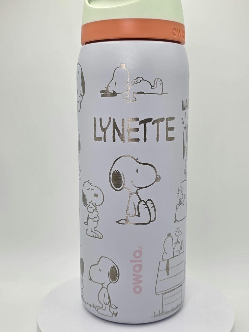 May include: A white insulated water bottle featuring gold Snoopy illustrations and the name "LYNETTE" in gold. The bottle has a light green and orange lid and the brand name "owala" in pink.
