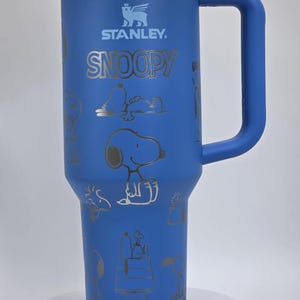 May include: Blue Stanley tumbler with a handle, featuring the Stanley logo and the word "SNOOPY" in silver. The tumbler is adorned with silver illustrations of Snoopy in various poses. The tumbler has a clear lid.