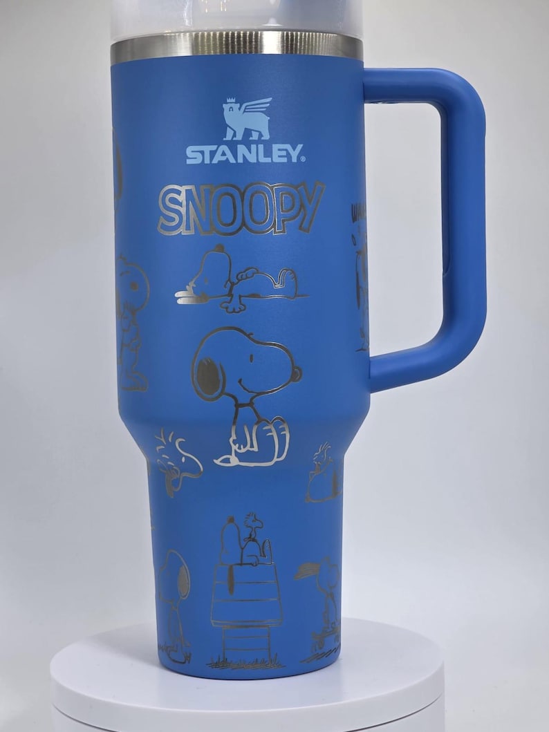 May include: Blue Stanley tumbler with a handle, featuring the Stanley logo and the word "SNOOPY" in silver. The tumbler is adorned with silver illustrations of Snoopy in various poses. The tumbler has a clear lid.