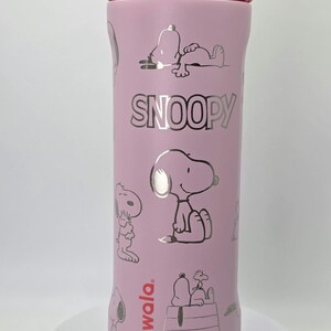 May include: A pink Owala water bottle featuring Snoopy illustrations. The bottle has a purple lid and a red band. The word "SNOOPY" is printed in silver. The bottle has a variety of Snoopy images.