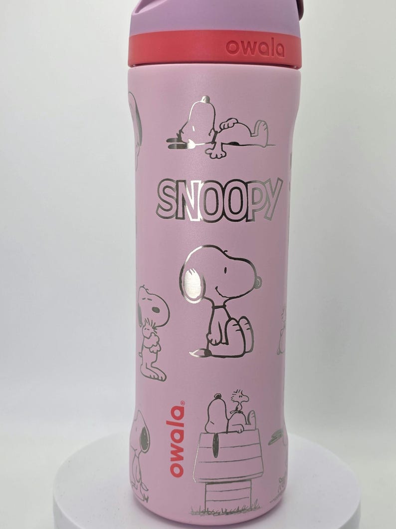 May include: A pink Owala water bottle featuring Snoopy illustrations. The bottle has a purple lid and a red band. The word "SNOOPY" is printed in silver. The bottle has a variety of Snoopy images.