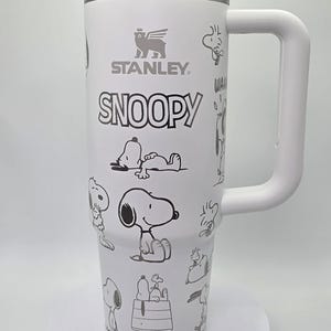 May include: White insulated tumbler with a handle, featuring the "STANLEY" and "SNOOPY" logos, along with various black and white Snoopy character illustrations. The tumbler has a silver rim and a white lid.