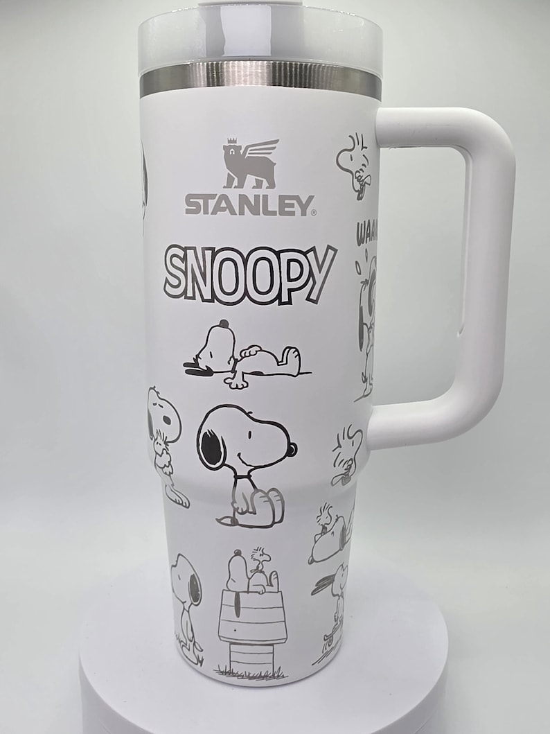 May include: White insulated tumbler with a handle, featuring the "STANLEY" and "SNOOPY" logos, along with various black and white Snoopy character illustrations. The tumbler has a silver rim and a white lid.