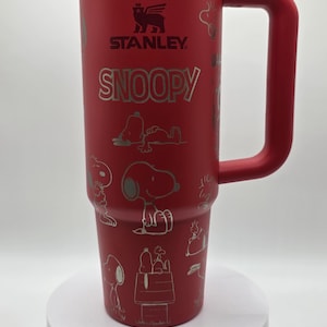 May include: A red Stanley travel mug with a large handle, featuring the Snoopy character in silver. The mug has the Stanley logo and Snoopy's name in silver. The design includes various Snoopy illustrations.
