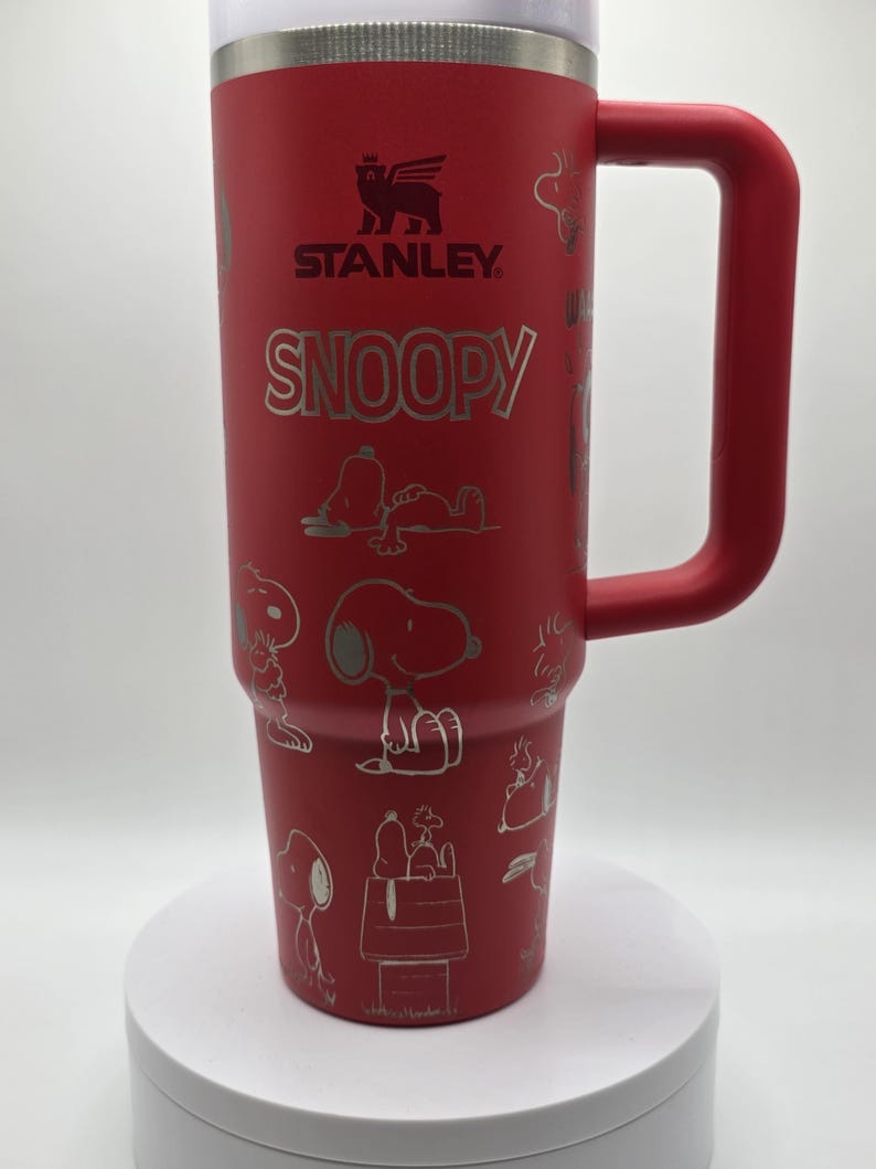 May include: A red Stanley travel mug with a large handle, featuring the Snoopy character in silver. The mug has the Stanley logo and Snoopy's name in silver. The design includes various Snoopy illustrations.