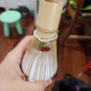 Op de afbeelding: Een bamboe matcha klopper met een wit en gouden keramische basis. Een parel- en kralenarmband omcirkelt de handgreep van de klopper, versierd met een bedel van twee rode kersen. De klopper wordt in een hand gehouden.