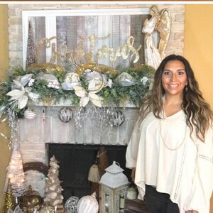 Pu&ograve; includere: Un camino in mattoni bianchi decorato con una ghirlanda natalizia argentata e dorata, un cartello dorato con la scritta "Natale" e una statuetta di angelo bianca. Una donna &egrave; in piedi davanti al camino con una blusa bianca e una collana di perle.