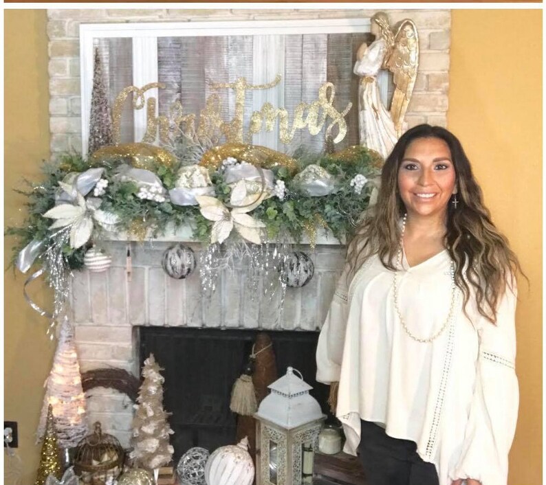 Pu&ograve; includere: Un camino in mattoni bianchi decorato con una ghirlanda natalizia argentata e dorata, un cartello dorato con la scritta "Natale" e una statuetta di angelo bianca. Una donna &egrave; in piedi davanti al camino con una blusa bianca e una collana di perle.