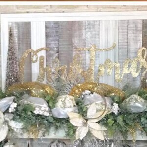 Pu&ograve; includere: Un cartello di Natale con glitter dorati con la parola "Natale" su uno sfondo di legno bianco. Il cartello &egrave; decorato con una ghirlanda di verde, fiori bianchi e glitter dorati.