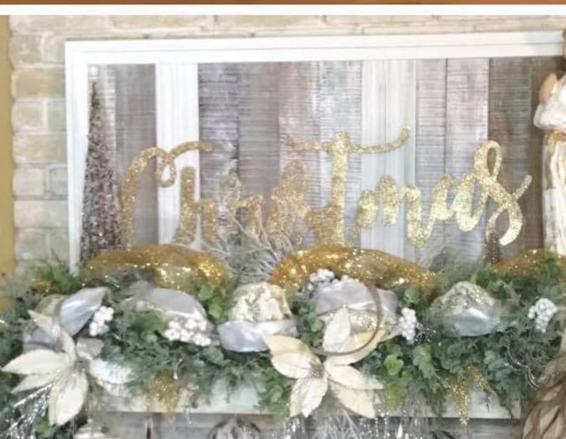 Pu&ograve; includere: Un cartello di Natale con glitter dorati con la parola "Natale" su uno sfondo di legno bianco. Il cartello &egrave; decorato con una ghirlanda di verde, fiori bianchi e glitter dorati.