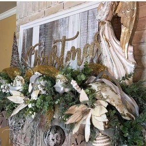 Pu&ograve; includere: Un cartello "Christmas" con glitter dorati &egrave; appeso sopra un camino decorato con verde, stelle di Natale bianche e nastro dorato. Una statuetta di angelo bianca con ali dorate si trova a destra del cartello.