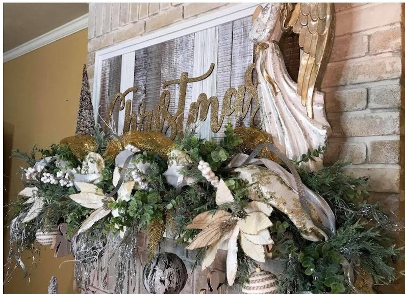 Pu&ograve; includere: Un cartello "Christmas" con glitter dorati &egrave; appeso sopra un camino decorato con verde, stelle di Natale bianche e nastro dorato. Una statuetta di angelo bianca con ali dorate si trova a destra del cartello.