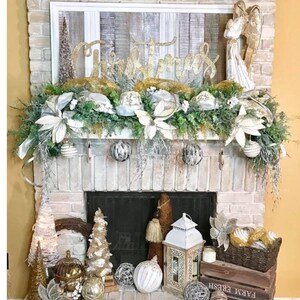 Pu&ograve; includere: Un camino in mattoni bianchi decorato per Natale con un cartello in legno bianco con la parola "Natale" in lettere dorate glitter, una statuetta di angelo bianco, verde, ornamenti bianchi e dorati e una lanterna bianca.