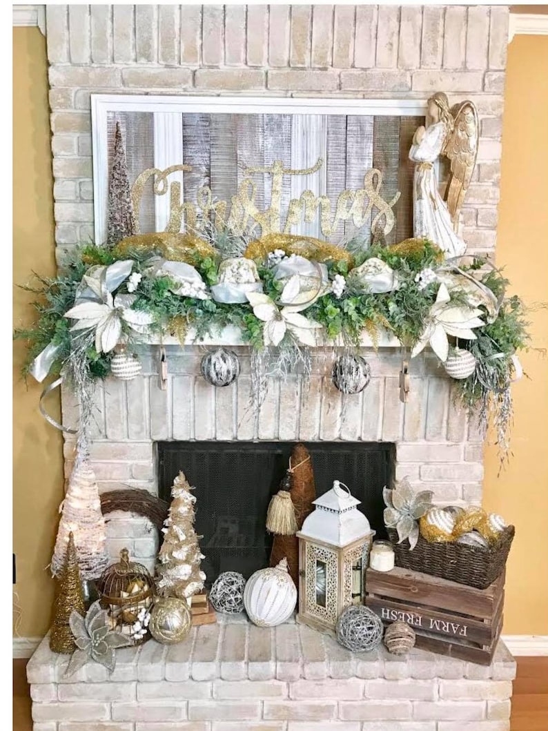 Pu&ograve; includere: Un camino in mattoni bianchi decorato per Natale con un cartello in legno bianco con la parola "Natale" in lettere dorate glitter, una statuetta di angelo bianco, verde, ornamenti bianchi e dorati e una lanterna bianca.