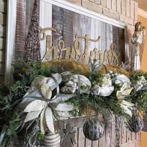 Pu&ograve; includere: Un cartello in legno sbiancato con la parola "Natale" in lettere dorate glitter, decorato con una ghirlanda di verde, stelle di Natale bianche e ornamenti argentati. La ghirlanda &egrave; drappeggiata su un camino in mattoni.