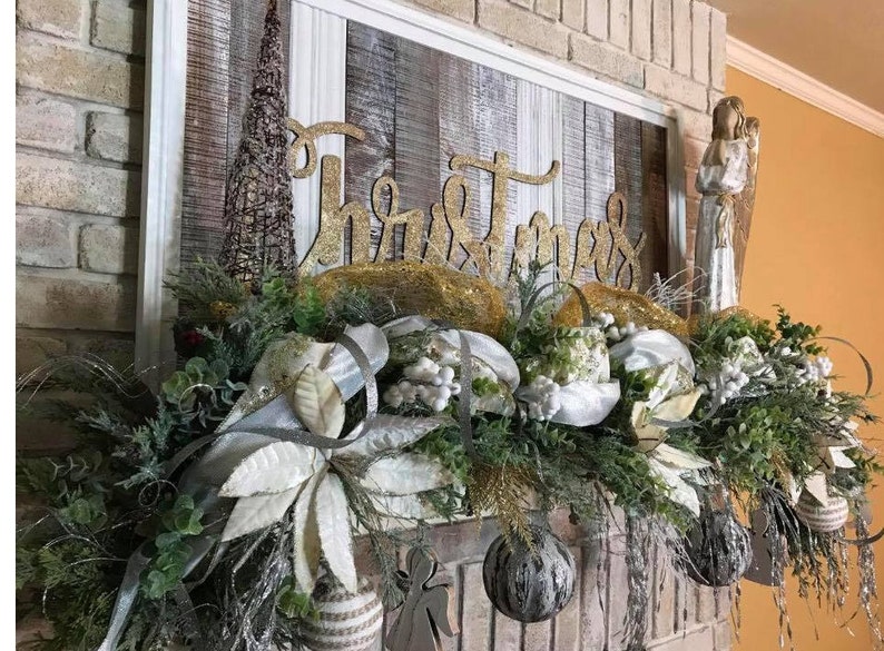 Pu&ograve; includere: Un cartello in legno sbiancato con la parola "Natale" in lettere dorate glitter, decorato con una ghirlanda di verde, stelle di Natale bianche e ornamenti argentati. La ghirlanda &egrave; drappeggiata su un camino in mattoni.