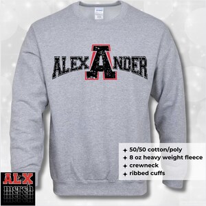 May include: A light gray crewneck sweatshirt with the name "ALEXANDER" in black distressed lettering with a red outline. The sweatshirt has ribbed cuffs and is made of 50/50 cotton/poly 8 oz heavy weight fleece.