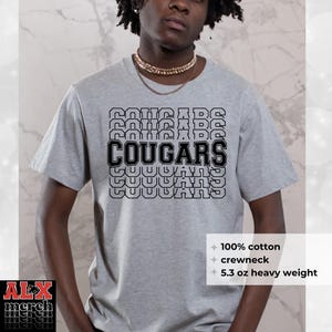 May include: A gray crewneck t-shirt with the word "COUGARS" in a stacked design. The shirt is made of 100% cotton and is 5.3 oz heavy weight. The shirt has the text "ALX merch" in the bottom left corner.