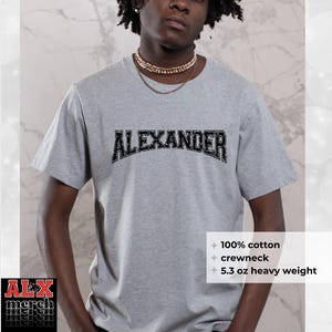 May include: A gray crewneck t-shirt with the name "ALEXANDER" in a black, distressed font across the chest. The shirt is made of 100% cotton and is 5.3 oz heavy weight. The ALX merch logo is visible in the bottom left corner.