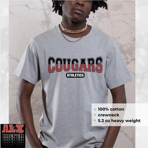 May include: A gray crewneck t-shirt with the word "COUGARS" in a black and red collegiate font. The word "ATHLETICS" is below the main text. The shirt is made of 100% cotton and is 5.3 oz heavy weight.