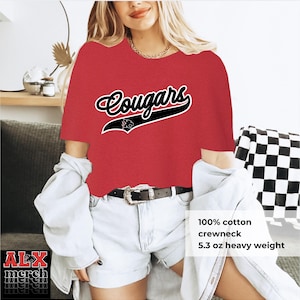 May include: Red crewneck t-shirt with the word "Cougars" in a script font with a black outline and a black graphic. The shirt is made of 100% cotton and is 5.3 oz heavy weight.