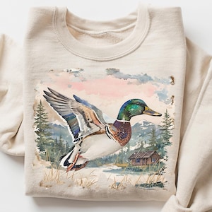 Pode incluir: Camisola bege com uma pintura em aquarela de um pato-real voando sobre uma paisagem com uma pequena cabana e árvores. O texto "CRAZY DEALS" está no canto superior direito.