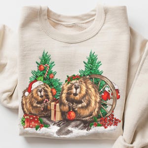 May include: Beige sweatshirt featuring a festive graphic of two beavers with Christmas trees, ornaments, and gifts. The image includes the text "CRAZY DEALS".