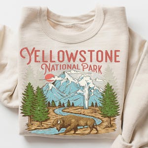 May include: Beige sweatshirt with a graphic of Yellowstone National Park. The design includes mountains, trees, a bison, and the text "Yellowstone National Park" in red. The sleeves are rolled up.