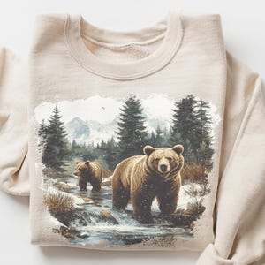 May include: Beige sweatshirt featuring a forest scene with two brown bears in a stream, surrounded by trees and mountains. The sweatshirt has long sleeves and a crew neck. The words "CRAZY DEALS" are in the upper right corner.