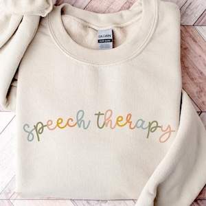 May include: A cream-colored sweatshirt with the words "speech therapy" in a colorful, cursive font. The sweatshirt has a crew neck and long sleeves. The background is a wooden surface with a chevron pattern.