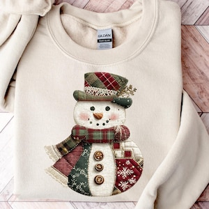 Puede incluir: Sudadera de color crema con un diseño caprichoso de muñeco de nieve. El muñeco de nieve lleva un sombrero a cuadros, una bufanda y ropa de estilo patchwork con detalles de botones. El diseño incorpora colores rojo, verde y marrón, perfecto para el invierno o las fiestas.