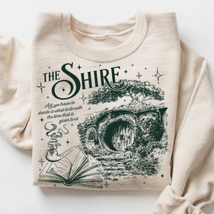 May include: Cream-colored sweatshirt featuring a green graphic of a hobbit hole with the text "The Shire." The design includes an open book and a smoking pipe. The sleeves are rolled up.