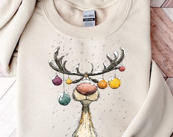 May include: A cream-coloured crew neck sweatshirt with a reindeer illustration. The reindeer has antlers decorated with baubles in various colours. The design includes falling snowflakes, creating a festive winter theme.