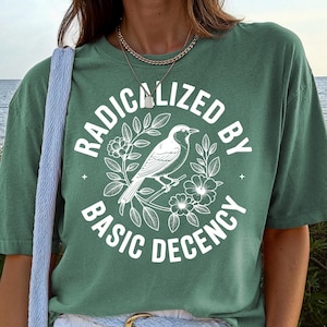 以下が含まれることがあります： 「RADICALIZED BY BASIC DECENCY」の文字と鳥と花のイラストが描かれたグリーンのTシャツ。白いショートパンツ、青いベルト、青いショルダーバッグとコーディネートされています。ゴールドのブレスレットとネックレスも着用しています。