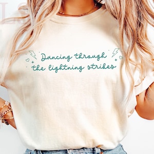 Pode incluir: Camiseta creme com a frase "Dancing through the lightning strikes" em verde-azulado. A camiseta é combinada com jeans azul claro. A estética geral é casual e descontraída.