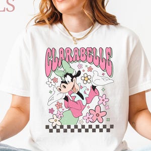 May include: White t-shirt with a cartoon Clarabelle Cow design in pink and green, surrounded by flowers. The word "CLARABELLE" is in pink bubble letters. The words "CRAZY DEALS" are in the upper left corner.