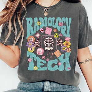 May include: Dark gray t-shirt with the words "Radiology Tech" in a retro font, surrounded by colorful flowers and medical-themed graphics. The shirt is worn with blue jeans and white sneakers.