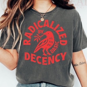 May include: Dark grey t-shirt featuring the red text "RADICALIZED BY BASIC DECENCY" and a red bird graphic. The shirt is worn with blue jeans and white trainers. A necklace and bracelets are also visible.