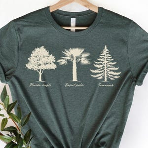 Può includere: T-shirt verde scuro con tre illustrazioni di alberi: acero della Florida, palma del deserto e larice, ciascuno etichettato sotto. La maglietta è esposta su una gruccia di legno su sfondo bianco. L'angolo in basso a destra della maglietta ha il testo "CRAZY DEALS".