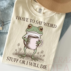 Puede incluir: Camiseta color crema con un gráfico de una rana verde sosteniendo una taza de café, con el texto "I HAVE TO SAY WEIRD STUFF OR I WILL DIE." La camiseta se combina con vaqueros azul claro, un sombrero beige y un collar.