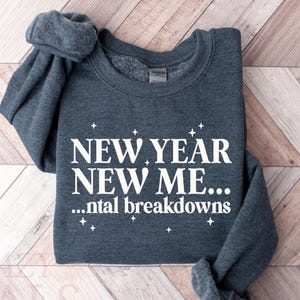 Puede incluir: Una sudadera gris oscuro con el texto blanco "NEW YEAR NEW ME... ...ntal breakdowns". La prenda está doblada sobre una superficie de madera marrón claro.