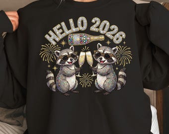 Puede incluir: Sudadera negra con un diseño de dos mapaches brindando con copas de champán. El diseño incluye el texto "HELLO 2026", una botella de champán, fuegos artificiales y detalles dorados. Los mapaches llevan gafas de sol.