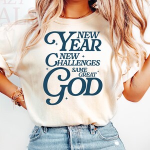 May include: Cream-coloured t-shirt with the text "New Year, New Challenges, Same Great God" in navy blue. The shirt is paired with light blue denim jeans. The overall style is casual and comfortable.