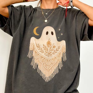 May include: Dark grey t-shirt featuring a whimsical ghost design. The ghost has a cream-colored body with a crescent moon and stars above it. The ghost's shawl is decorated with floral and fringe details. The shirt is a relaxed fit.