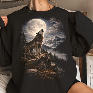 May include: A black crewneck sweatshirt featuring a graphic print of a wolf howling at a full moon. The scene depicts a rocky cliffside, a dense pine forest, and snow-capped mountains under a night sky with clouds, rendered in shades of grey and white.