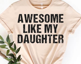 May include: A pale beige t-shirt with the phrase "AWESOME LIKE MY DAUGHTER" printed in bold, black letters. The shirt is displayed on a wooden hanger. Green foliage and a branch are positioned on the left. The words "CRAZY DEALS" are in the bottom right corner.