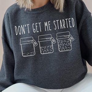 May include: Dark gray sweatshirt with the text "DON'T GET ME STARTED" above three jars with contents. The jars have tags hanging from them. The sweatshirt is worn with white pants.