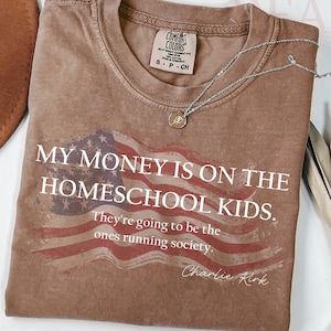 Pu&ograve; includere: Una maglietta marrone con la scritta "MY MONEY IS ON THE HOMESCHOOL KIDS. They're going to be the ones running society." e un disegno della bandiera americana. Una collana d'argento con un ciondolo &egrave; sulla maglietta.