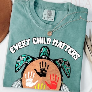 Op de afbeelding: Een turquoisekleurig T-shirt met de witte tekst "EVERY CHILD MATTERS". Het shirt heeft een schildpadontwerp met kleurrijke handafdrukken. Een zilveren ketting met een hanger is over het shirt gedrapeerd.